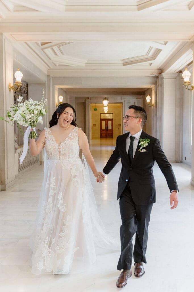 Couple holding hands and walking on the third floor of San Francisco City Hall with bouquet